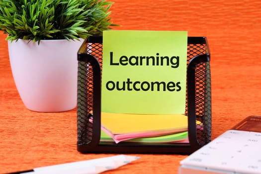 Learning Outcome write on a green sticker next to office supplies on an orange background