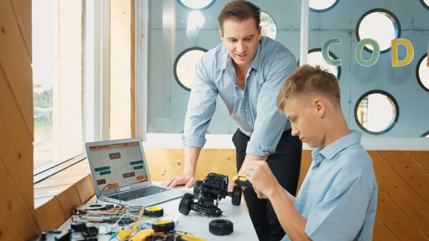 Caucasian teacher help student fixing car model and giving advise for programing system or coding prompt while highschool boy listen and use equipment to repair robot at STEM classroom. Edification.