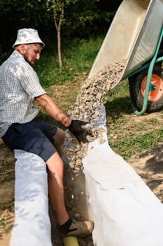 A man pours rubble crushed stone from a wheelbarrow into a trench. Drainage works for drainage of ground water around the field.
