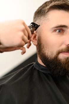 Barber shaves the contour of the oval line with a clipper on the clients head. A man with a beard gets a haircut in a barbershop chair.