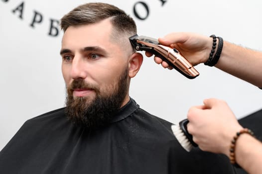 A barber cuts a man with a beard in a barber shop. Short haircut of the client with a clipper.