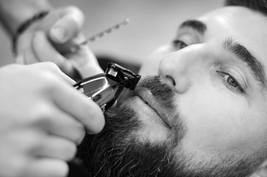 A barber trims a Caucasian client mustache with a trimmer.