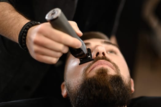A barber trims a Caucasian client mustache with a trimmer.