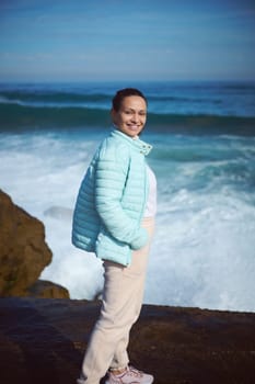 Happy smiling woman standing by ocean on the rocky cliff, looking at camera, enjoying the view of beautiful waves making white foam while crushing on the headland