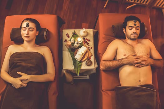 Top view couple enjoying relaxing anti-stress head massage with hot stone and pampering facial beauty skin recreation leisure in warm candle light ambient salon spa in resort or hotel. Quiescent