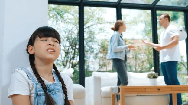 Stressed and unhappy young girl huddle in corner, cover her ears blocking sound of her parent arguing in background. Domestic violence at home and traumatic childhood develop to depression. Synchronos