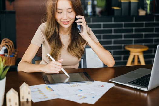 Freelance Asian business woman sitting at cafe call mobile phone talking and asking about something, smiling female working laptop computer and take note on digital tablet at home office