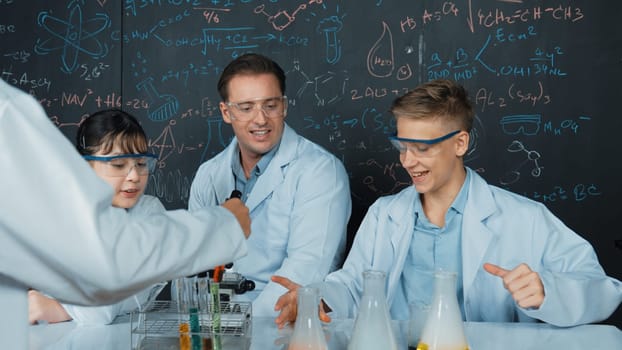 Creative teacher pointing chemistry at blackboard while talking to highschool boy at table with microscope and test tube with colored liquid. Young student wearing lab coat in STEM class. Edification.