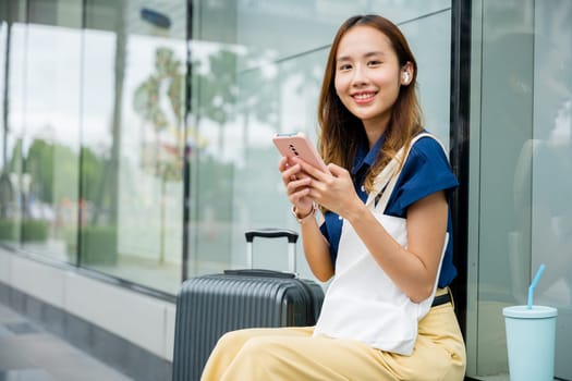 A young woman sitting with her suitcase, using her mobile phone to stay connected and multitasking while waiting for her flight at the airport. She is modern and stylish.