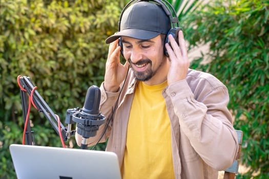 A man with a beard and moustache wearing a cap and headphones is singing and recording podcast into a microphone while using a laptop outdoor. He looks happy and relaxed, enjoying a leisurely musical event
