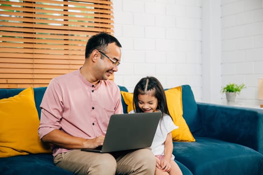 A smiling Asian father works from home on his laptop as his daughter learns on a computer for e-learning. In their modern living room they share moments of togetherness bonding and happiness.