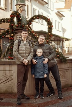 Joyful Family Portrait: Father and Two Sons by Festive Vintage Fountain. Capture the essence of familial happiness with this heartwarming image featuring a handsome father with his two sons standing against the backdrop of a festively decorated vintage fountain. The image beautifully blends the joy of the holiday season with the timeless charm of family togetherness. The father is 44 years old, the elder son is 17, and the younger one is 8, creating a memorable and multigenerational festive scene.