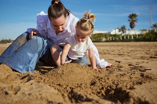 Happy woman and her cute little child girl enjoying time together, sitting on the sandy beach on warm sunny day, building castles while walking together outdoor