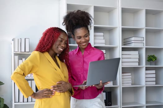 Two African American businesswomen using laptop and clipboards stand talking, giving advice on work and planning accounting and finance work together in the office..