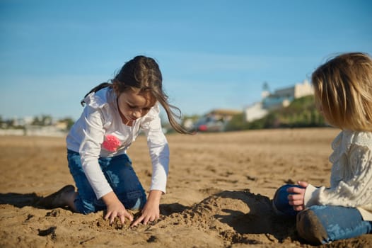 Two Caucasian adorable little kids girls playing together on the beach, building sandy castle on warm sunny day