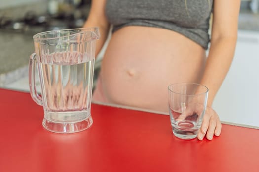 Embracing the vital benefits of water during pregnancy, a pregnant woman stands in the kitchen with a glass, highlighting hydration's crucial role in maternal well-being.