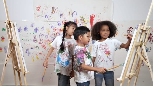 Diverse student painted or draw canvas at stained wall in art lesson. Asian girl wearing white shirt with stained color while standing at stained wall with hand print. Creative activity. Erudition.