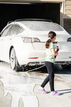 A young girl enthusiastically assists in washing the family's electric car in their suburban driveway.