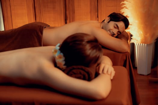 Caucasian couple customer enjoying relaxing anti-stress spa massage and pampering with beauty skin recreation leisure in warm candle lighting ambient salon spa at luxury resort or hotel. Quiescent