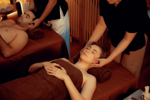 Couple customer enjoying relaxing anti-stress head massage and pampering facial beauty skin recreation leisure in warm candle lighting ambient salon spa in luxury resort or hotel. Quiescent