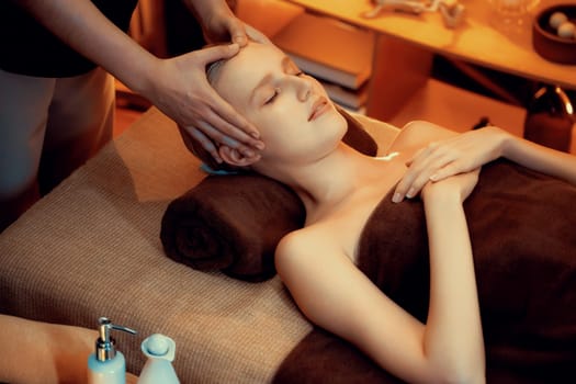 Caucasian woman enjoying relaxing anti-stress head massage and pampering facial beauty skin recreation leisure in warm candle lighting ambient salon spa in luxury resort or hotel. Quiescent