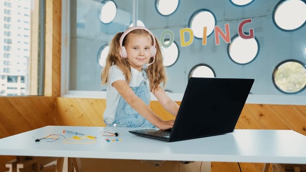 Little young cute girl using laptop to working or playing games. Student working on computer while looking and smiling at camera. Caucasian child attend online class by using headphone. Erudition.