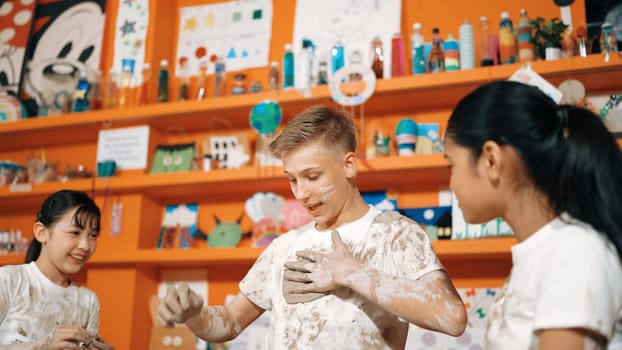 Smart caucasian teenager and happy friend with mixed races put clay on body in art lesson. Diverse highschool student playing and modeling cup of clay at workshop in creative activity. Edification.