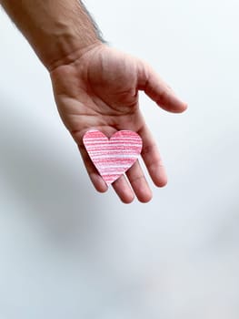 Mans hand holding striped pink paper heart against white background, symbolizing love and care, with copy space. Can be used for design and print related to childrens, love and relationships themes, charity and healthcare campaigns, emotional and mental well being content. High quality photo