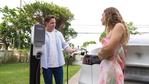 Happy and lovely couple with eco-friendly conscious recharging electric vehicle from EV charging station. EV car technology utilized as alternative transportation for future sustainability. Synchronos