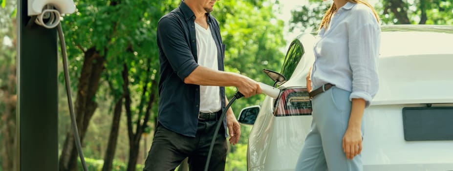 Focused EV car recharging electricity for battery on blurred background of lovey couple ruing their road trip travel by eco friendly electric car in national park and greenery forest on holiday. Exalt