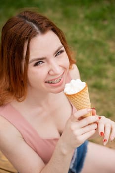 Young beautiful red-haired woman smiling with braces and eating an ice cream cone outdoors. Vertical photo