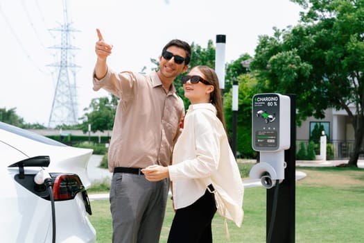 Young couple with coffee pay for electricity while recharge EV car battery at charging station connected to electrical power grid tower as electric industrial for eco friendly car travel. Expedient