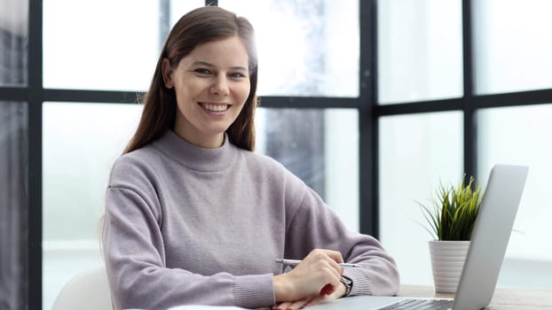 A woman in close-up at a desk in the office working and looking at the camera with a smile.
