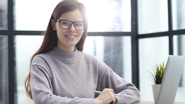 A woman with glasses in close-up at a desk in the office working and looking at the camera with a smile.