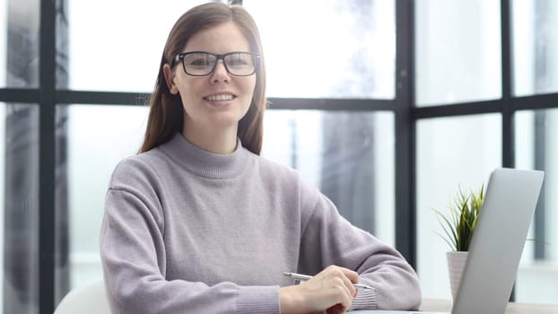 A woman with glasses in close-up at a desk in the office looking at the camera with a smile.