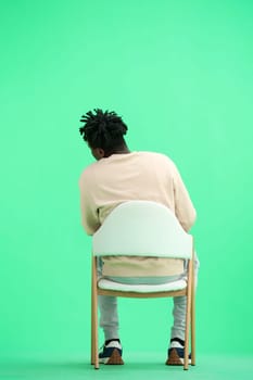 A man, on a green background, sitting on a chair.