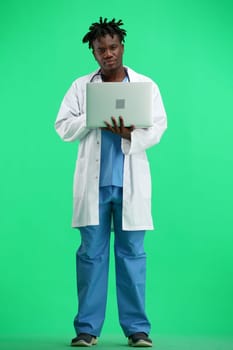 The doctor, in full height, on a green background, uses a laptop.