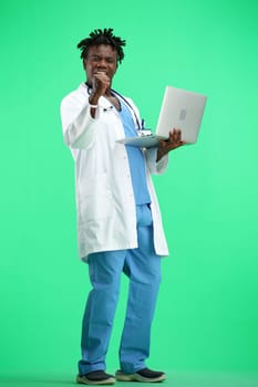 The doctor, in full height, on a green background, uses a laptop.