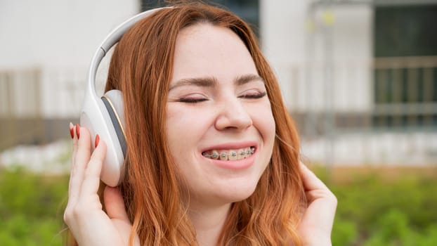 Portrait of a young red-haired woman with braces on her teeth listening to music on headphones outdoors