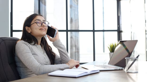A young woman in the office with a laptop talking on the phone.