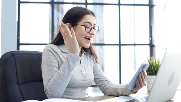 A young woman with glasses in the office is working on a laptop and talking on the phone.