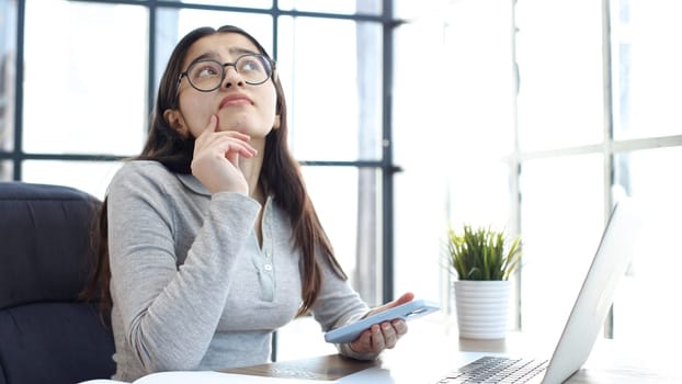 A young woman with glasses in the office is working on a laptop and talking on the phone.