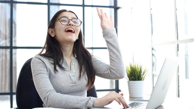 A young woman with glasses in the office waves to a colleague.
