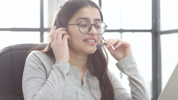 A young woman in close-up with glasses and headphones works in a call center.