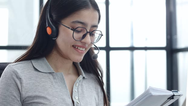 A young woman in close-up with glasses and headphones works in a call center.