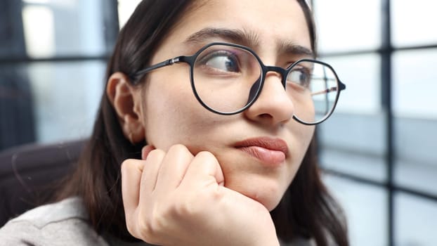 A young woman's face in close-up with glasses thinks.