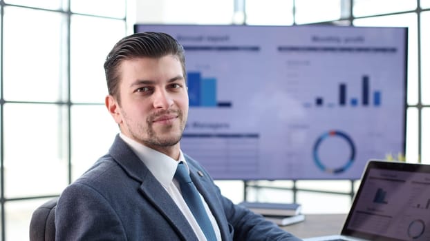 A businessman man in a close-up in a jacket with graphs on the background looks at the camera.