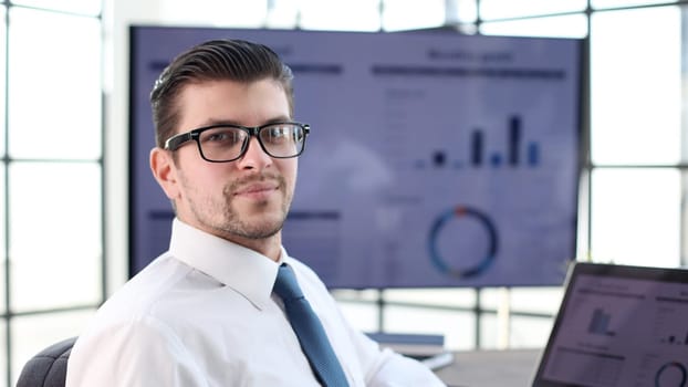 A businessman man in close-up in a shirt with graphs on the background looks at the camera.