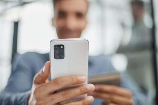 Formally dressed man sits on a sofa and enters a credit card number into a smartphone to pay online.