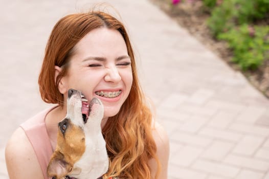 Dog jack russell terrier licks the owner in the face outdoors. Girl with braces on her teeth